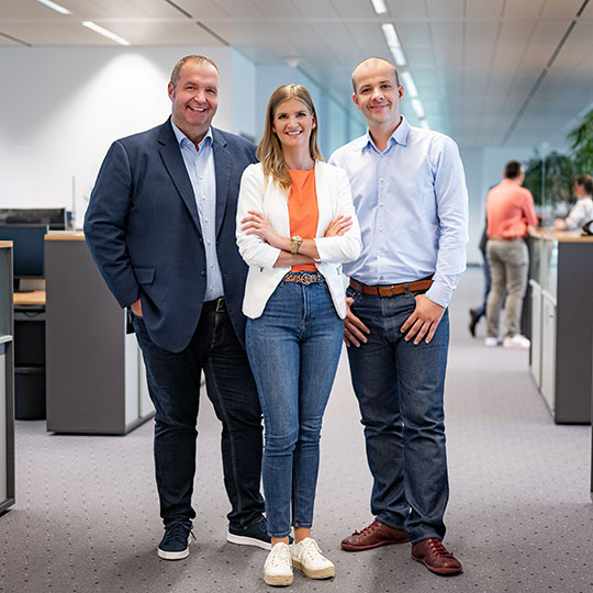 Mitarbeitergruppe aus drei Personen stehend im Büro mit Blick Richtung Kamera